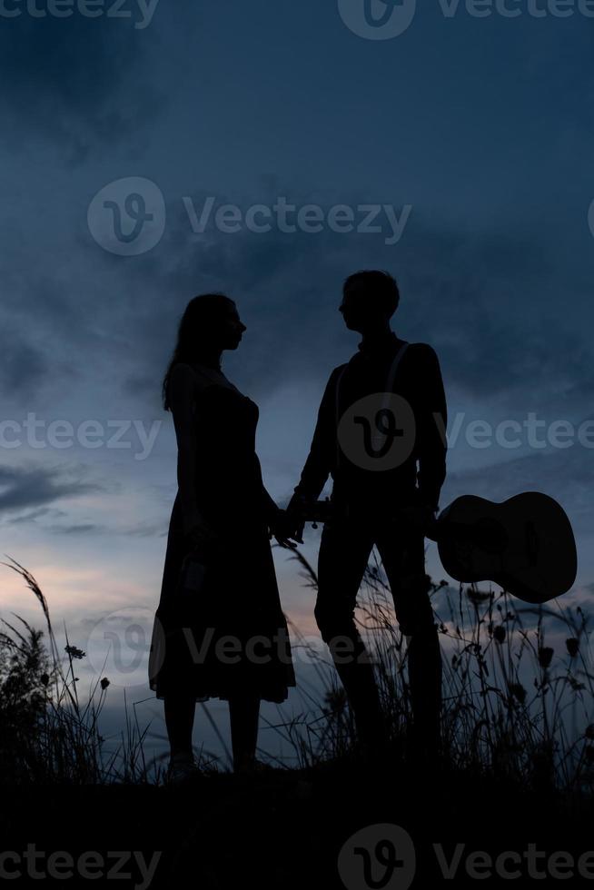 sagoma di una coppia innamorata ad un appuntamento. l'uomo tiene la mano della ragazza. il tizio in granchi ha una chitarra. girato al tramonto. foto