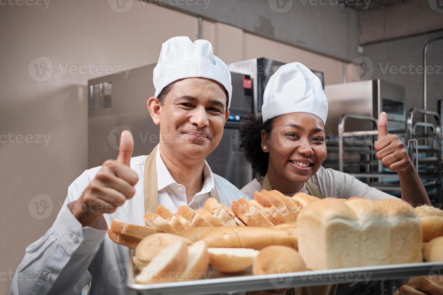 ritratto di chef professionisti in uniforme bianca guardando la fotocamera con un sorriso allegro e pollice in su con vassoio di pane in cucina. amico e partner di prodotti da forno e dell'occupazione quotidiana di prodotti da forno freschi. foto