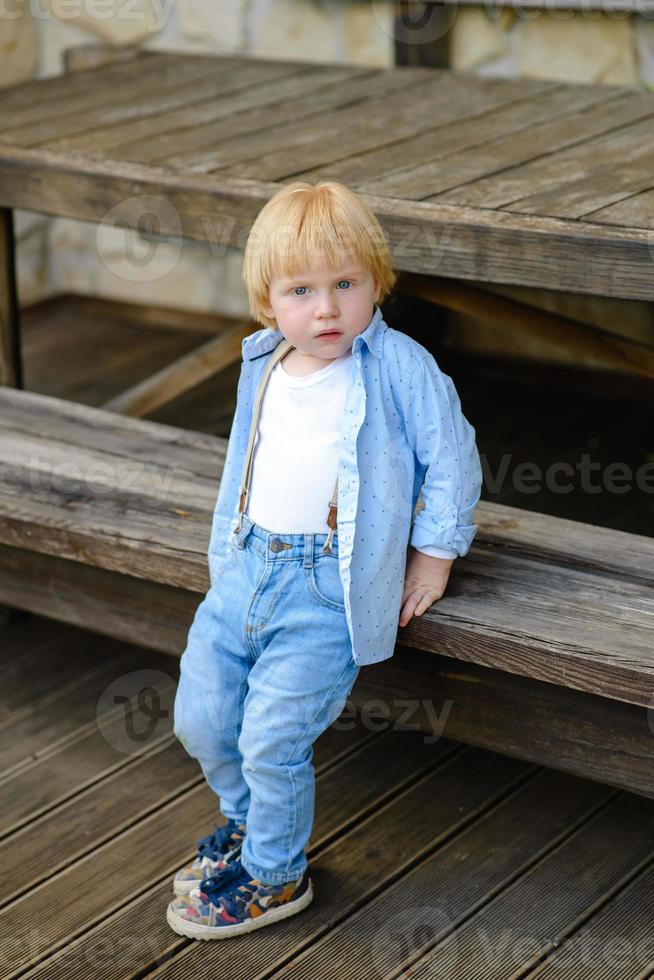 piccolo ragazzo biondo si siede su una panca di legno foto