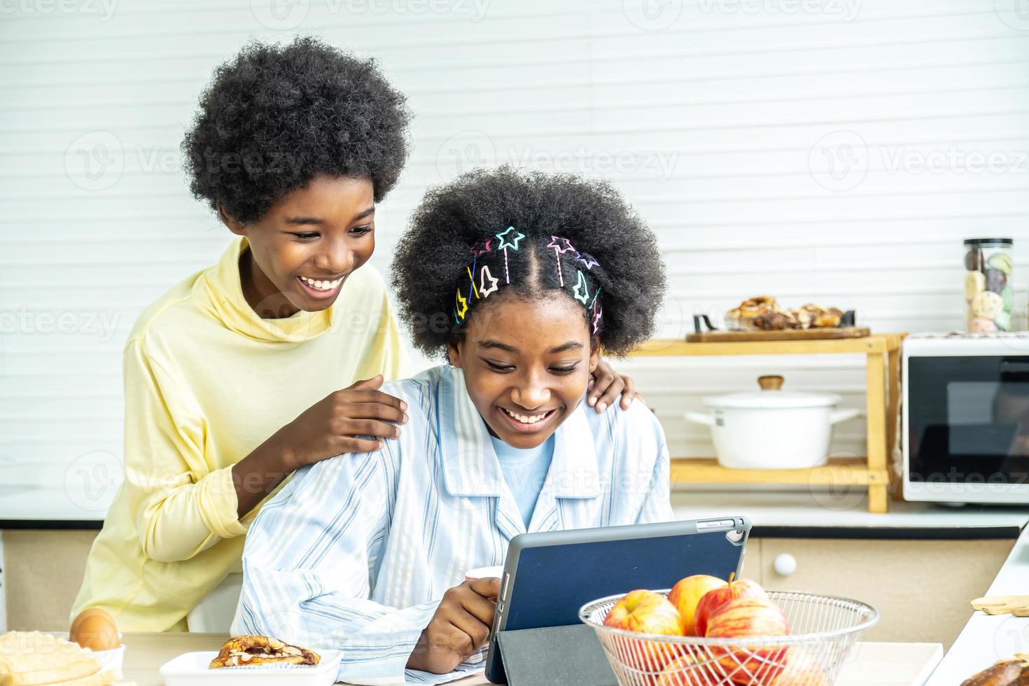 due bambini afroamericani felici in cucina mentre fanno colazione e tè o latte nella tazza inviano un messaggio o guardano film con il tablet, concetto di social network, messaggio, tecnologia foto