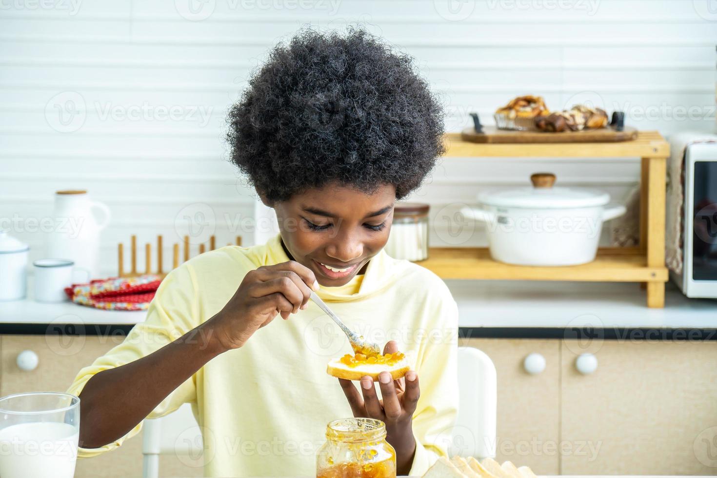 simpatico ragazzo afroamericano che sparge marmellata sul pane e mangia in cucina, lo studente si gode la colazione o spuntini di pane tostato con marmellata foto