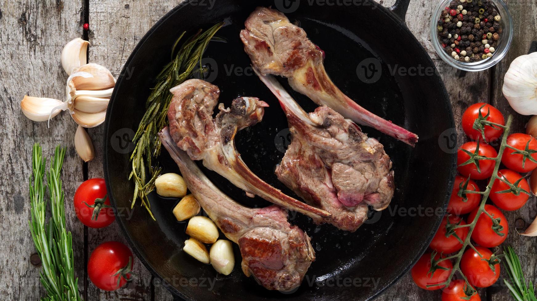 costolette di agnello arrosto con rosmarino, pomodori, aglio in padella su fondo di legno rustico scuro foto