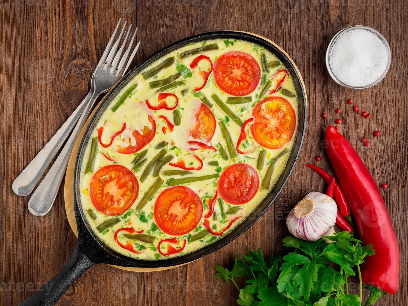 padella ovale in ghisa con deliziosa frittata calda con pomodori e peperoni. sfondo di struttura in legno marrone, primo piano, vista dall'alto. foto