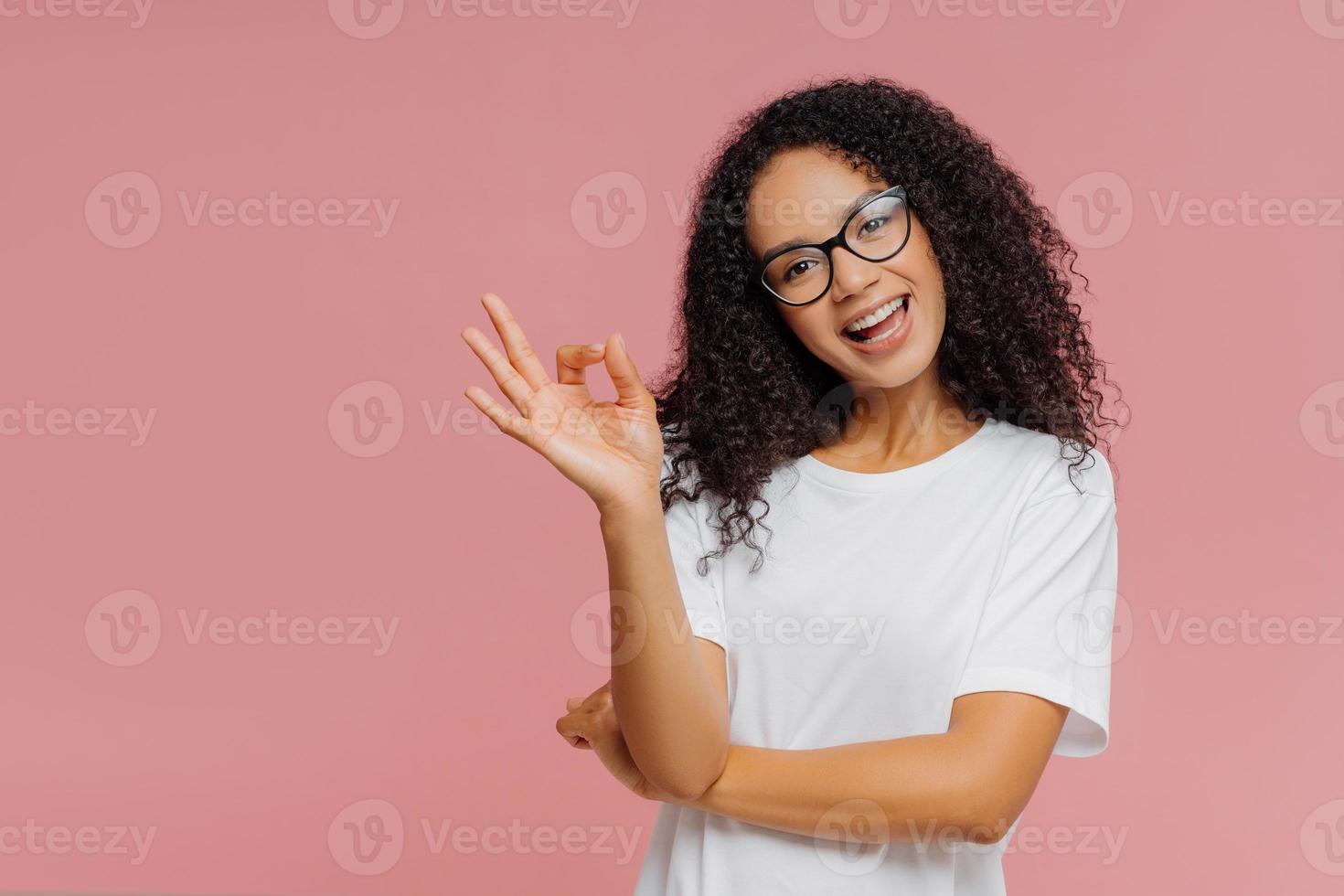 ottimista bella donna dalla pelle scura fa un gesto ok, inclina la testa, dimostra approvazione, è d'accordo con qualcosa, ha un'espressione felice, indossa occhiali e maglietta bianca casual, isolata sul muro rosa foto