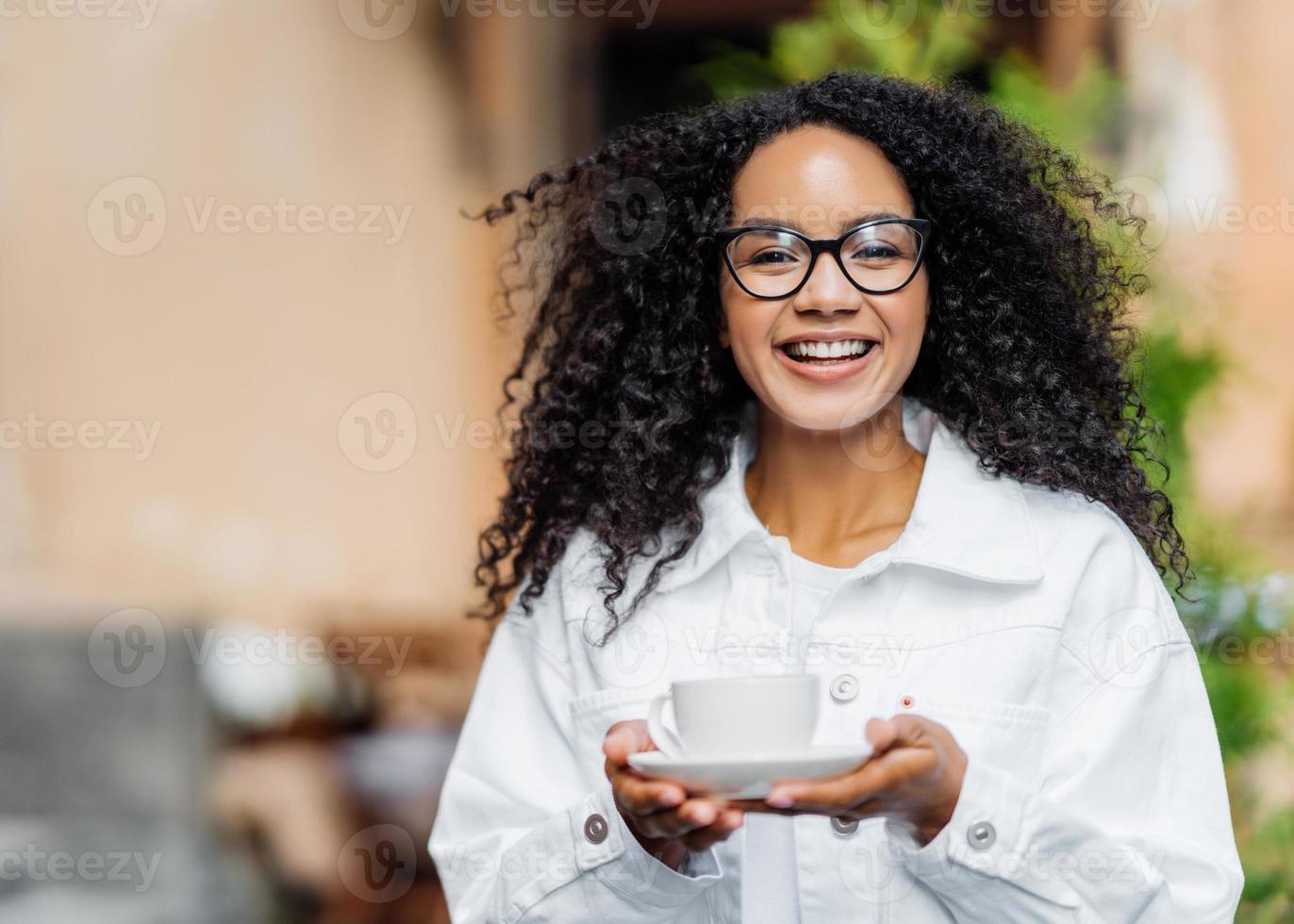 scatto all'aperto di una donna felice dalla pelle scura con i capelli crespi, indossa una giacca bianca, beve un tè caldo, passeggia dall'altra parte della strada, un'espressione felice. la donna afro soddisfatta gode del tempo libero durante il fine settimana foto