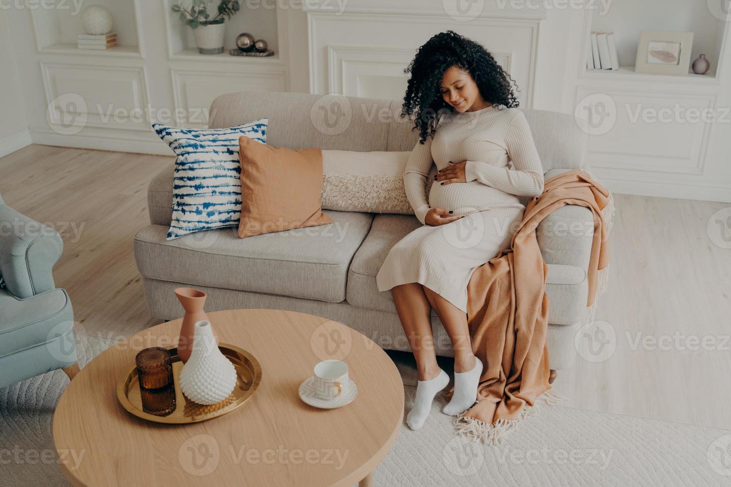 bella foto afro americana signora seduta sul divano accogliente e in attesa di un bambino, guardando in basso sulla sua pancia