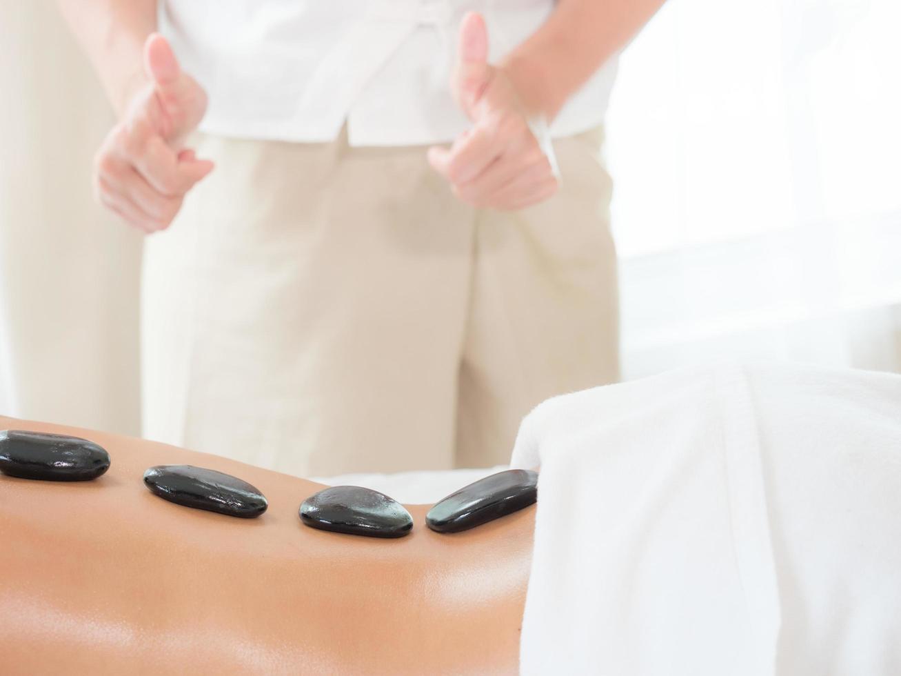 una massaggiatrice esperta in un salone termale mette pietre calde sulla schiena di una donna asiatica per alleviare la tensione dal lavoro foto