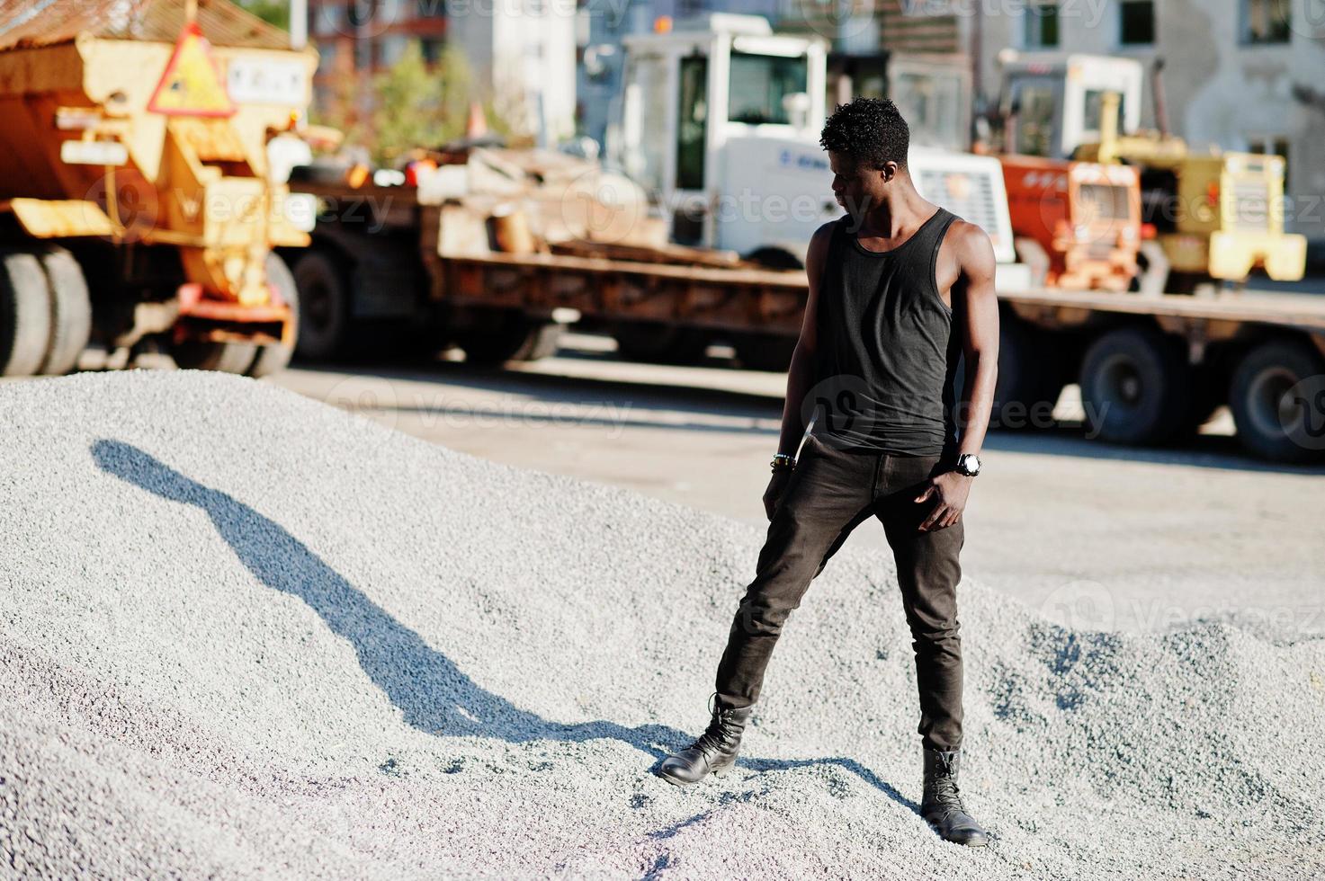 attraente ragazzo afroamericano nero in camicia muscolare nera in posa sul mucchio di ghiaia nella zona industriale. foto