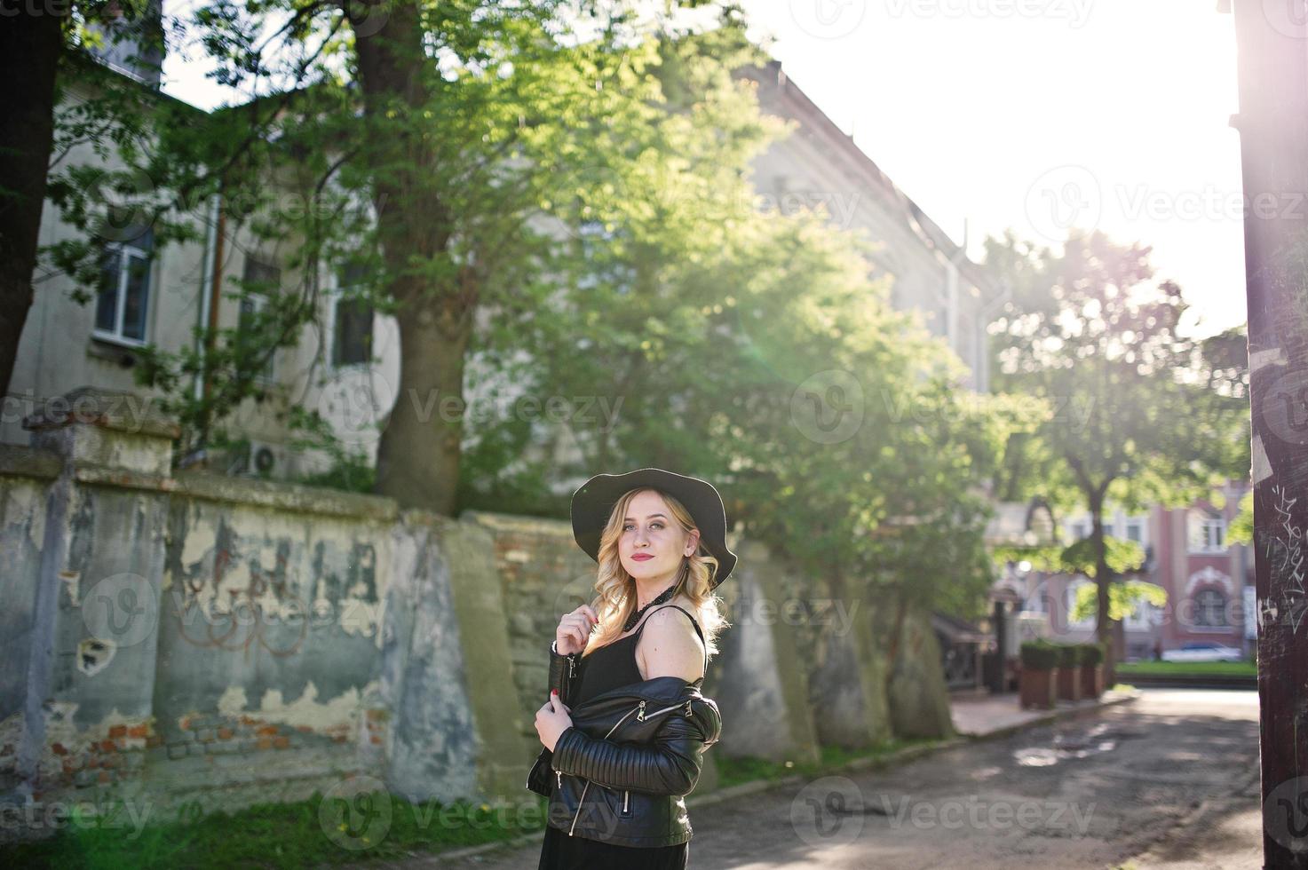 donna bionda su abito nero, giacca di pelle, collane e cappello in posa contro la strada della città al tramonto. foto