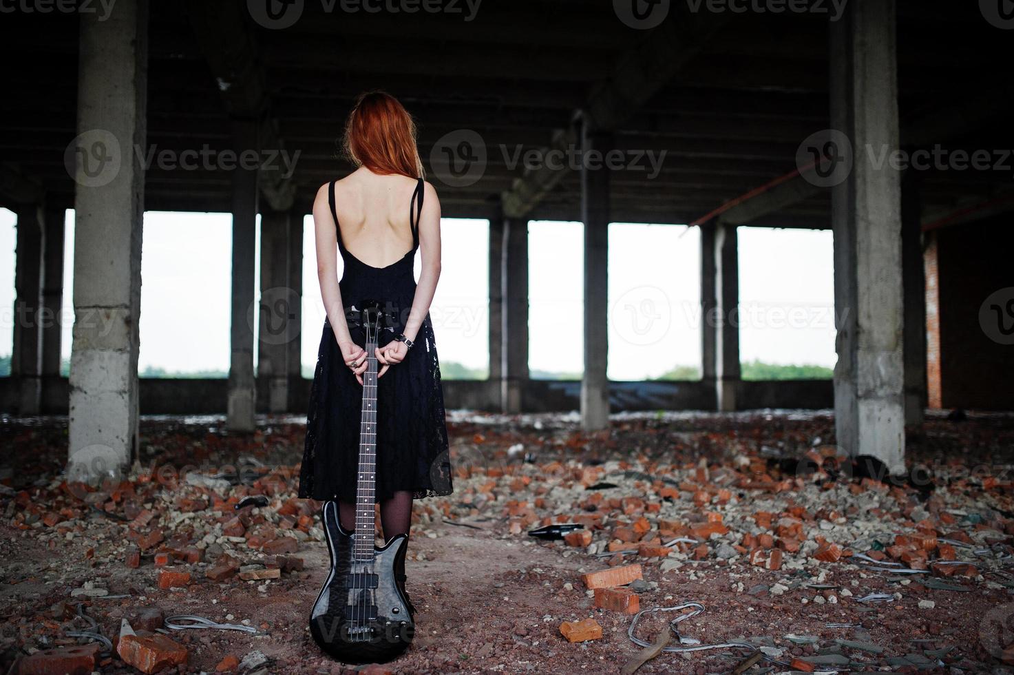 la ragazza punk dai capelli rossi indossa il nero con il basso in un luogo abbandonato. ritratto di musicista donna gotica. foto