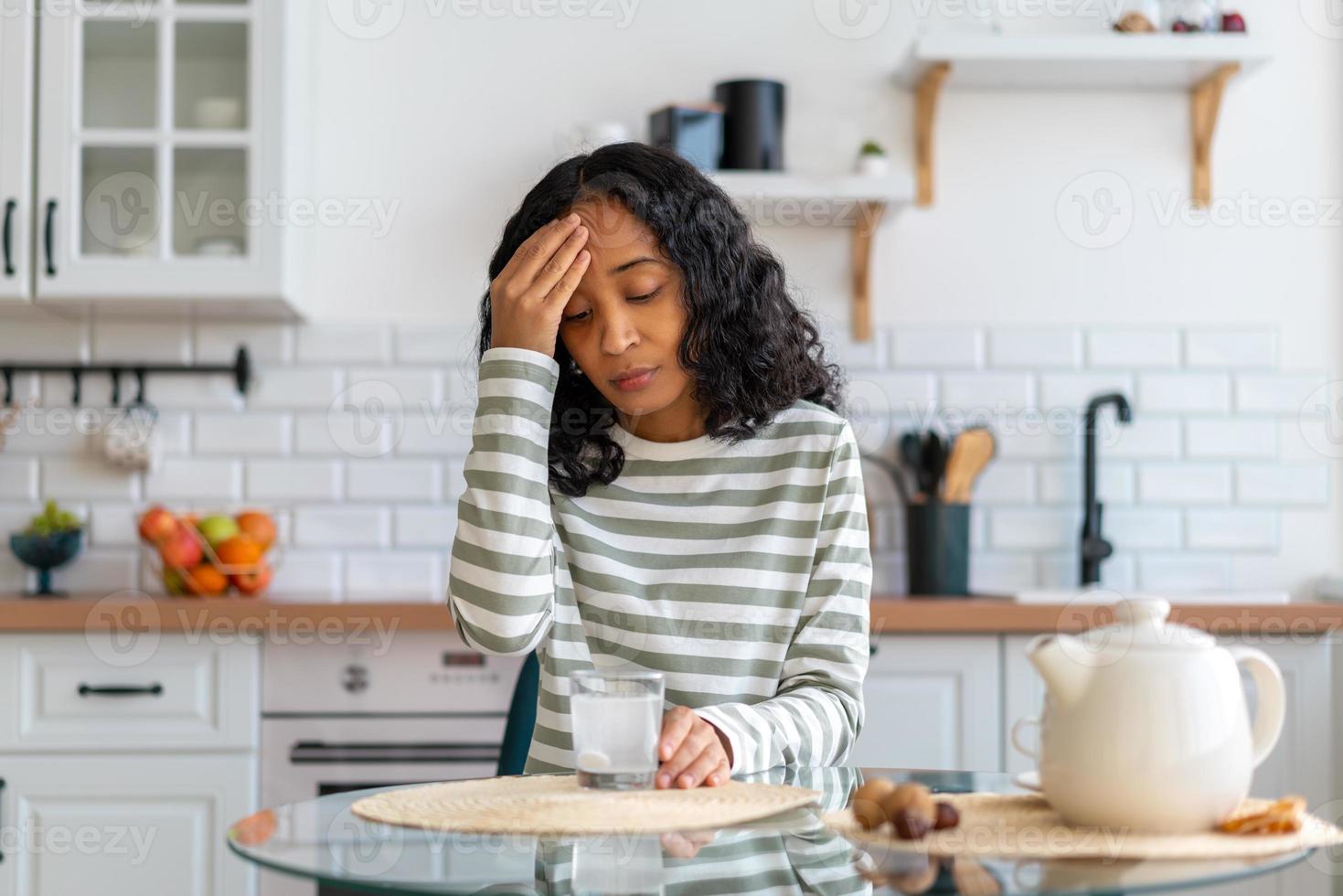 femmina afro-americana che si occupa di mal di testa. pillola dissolvente in un bicchiere d'acqua in cucina foto