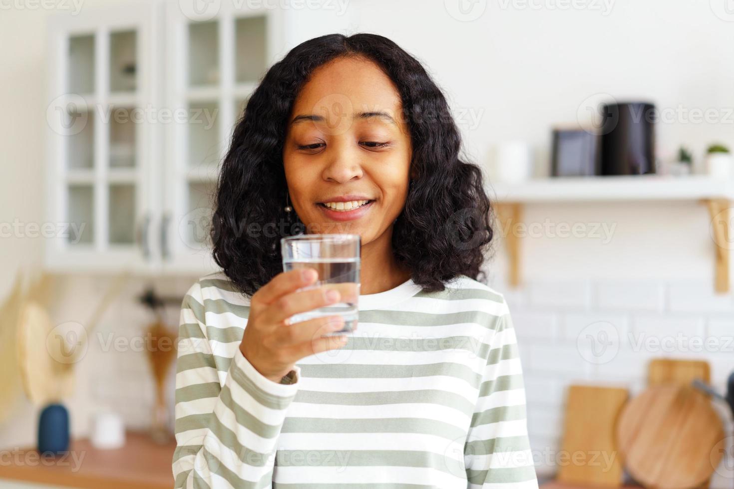 femmina afro-americana sorridente che gode di un bicchiere di acqua limpida mentre è in piedi in cucina foto