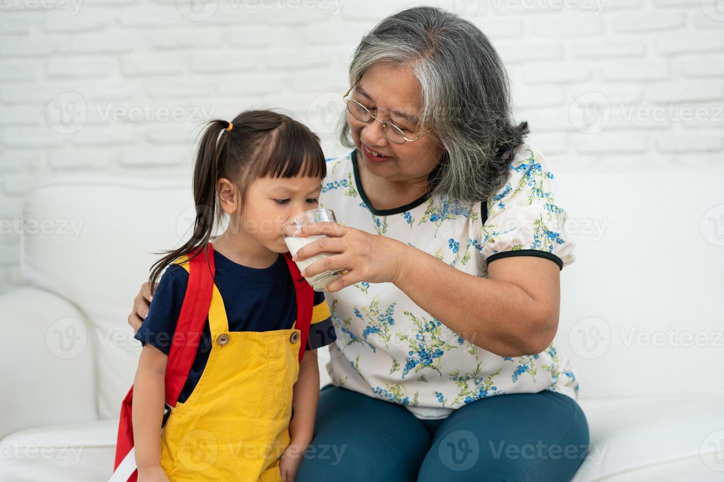 felice anziana nonna asiatica si siede accanto a sua nipote e nutre il latte fresco dal bicchiere per colazione a casa. concetto di una famiglia felice e si prende cura insieme, assistenza sanitaria in età prescolare foto