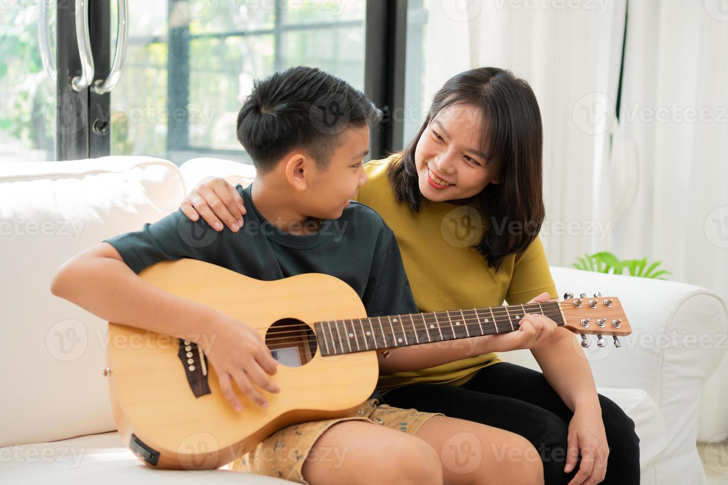 la madre asiatica abbraccia il figlio, il ragazzo asiatico che suona la chitarra e la madre abbraccia sul divano e si sente apprezzata e incoraggiata. concetto di famiglia felice, stile di vita di apprendimento e divertimento, legami familiari d'amore foto