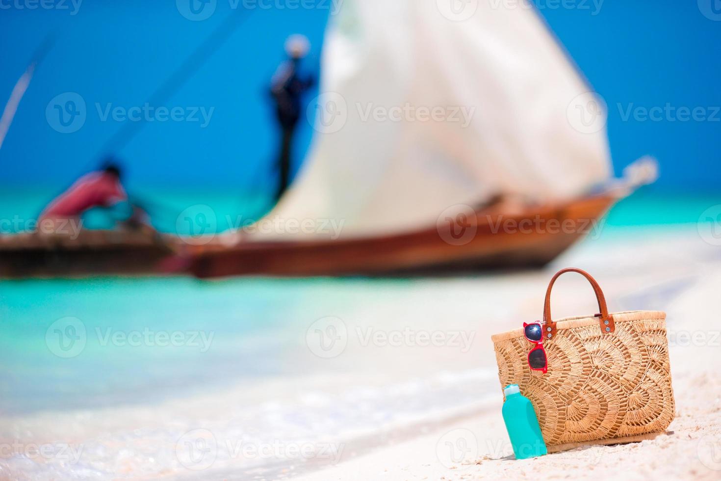 accessori da spiaggia - borsa di paglia, bottiglia di crema solare e occhiali da sole rossi sulla spiaggia foto
