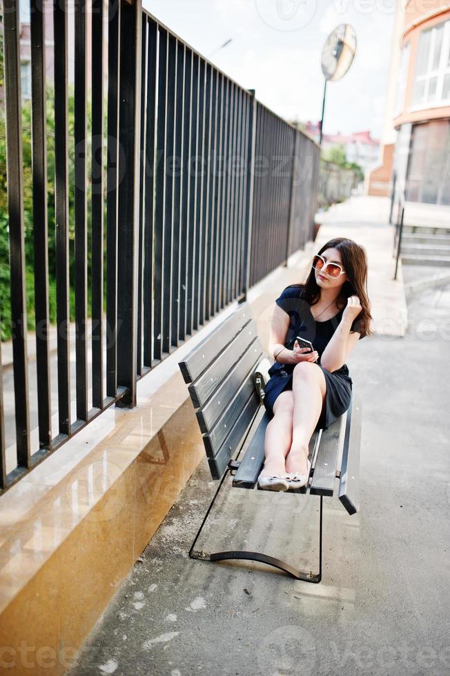 ragazza bruna in abito nero, occhiali da sole seduti su una panchina, ascoltando musica dalle cuffie e posando in una strada della città. foto