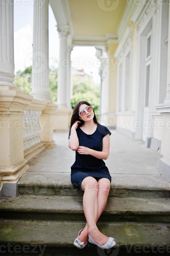 ragazza bruna in abito nero, occhiali da sole seduti sulle scale della vecchia casa d'epoca, in posa in una strada della città. foto