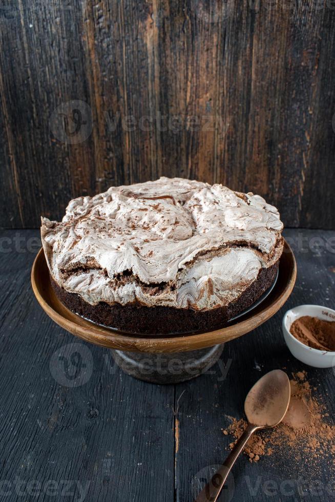 torta brownie al forno rotonda condita con meringa e vortici di cioccolato su un supporto per torta rustico foto