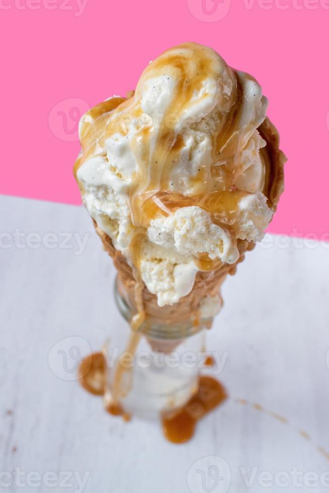 scioglimento di palline di gelato alla vaniglia con salsa al caramello gocciolante su un cono di cialda su uno sfondo rosa vivace e divertente foto