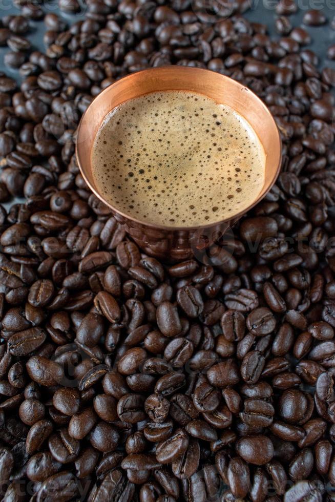 tazza di rame piena di caffè espresso al centro di chicchi di caffè crudo sparsi su un tavolo rustico foto