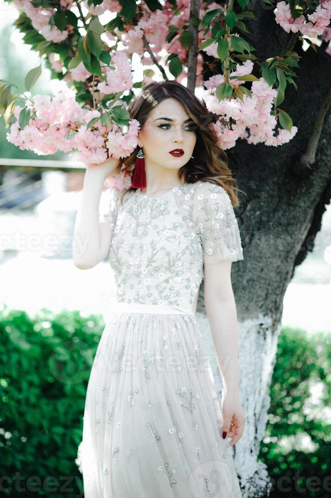 ragazza molto bella la sposa sotto un velo, abito da sposa beige vicino all'albero sakura sta guardando in basso, la natura foto