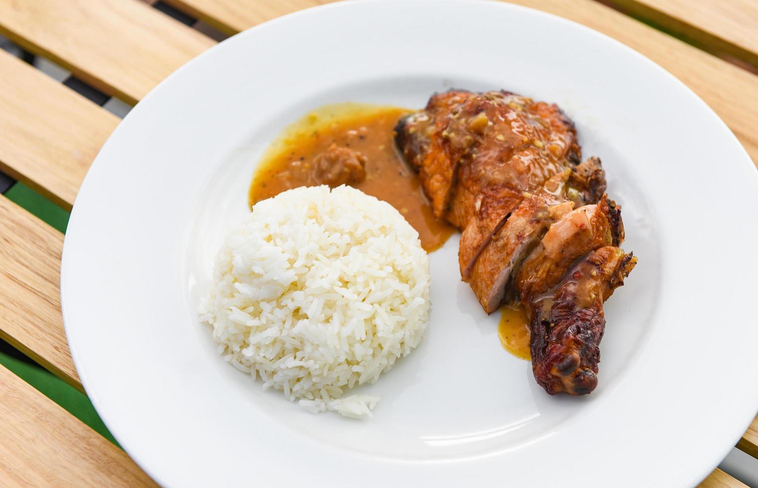 cibo di riso tailandese, riso bianco cotto e pollo alla griglia con salsa su piatto bianco e sfondo tavolo in legno, cosce di pollo barbecue speziate alla griglia foto