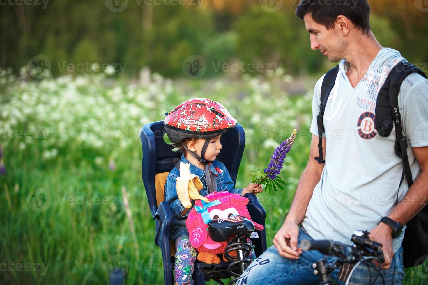 padre con una figlia piccola nel seggiolino di una bicicletta per bambini su una bicicletta per adulti. una ragazza con un casco protettivo, con una banana e uno zaino. passeggiata sportiva in famiglia, sicurezza. Kaluga, Russia, 30 maggio 2018 foto