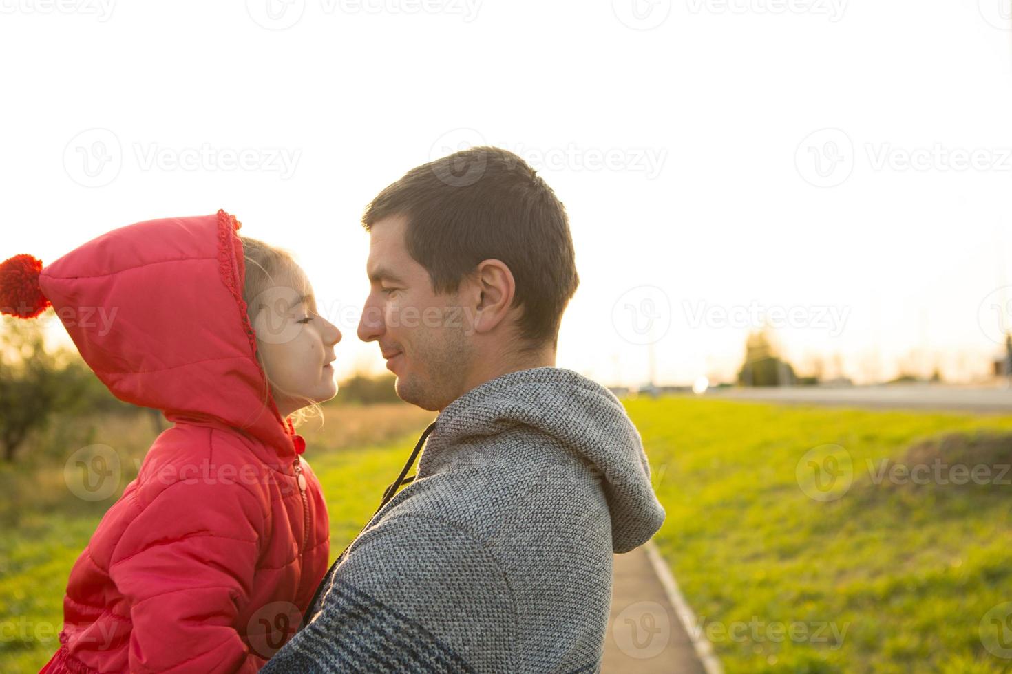 bambina in giacca rossa con cappuccio abbraccia e bacia suo padre, sorride, le tocca il naso. famiglia felice, emozioni dei bambini, festa del papà, raggi luminosi del sole, aspetto caucasico. spazio per il testo. foto