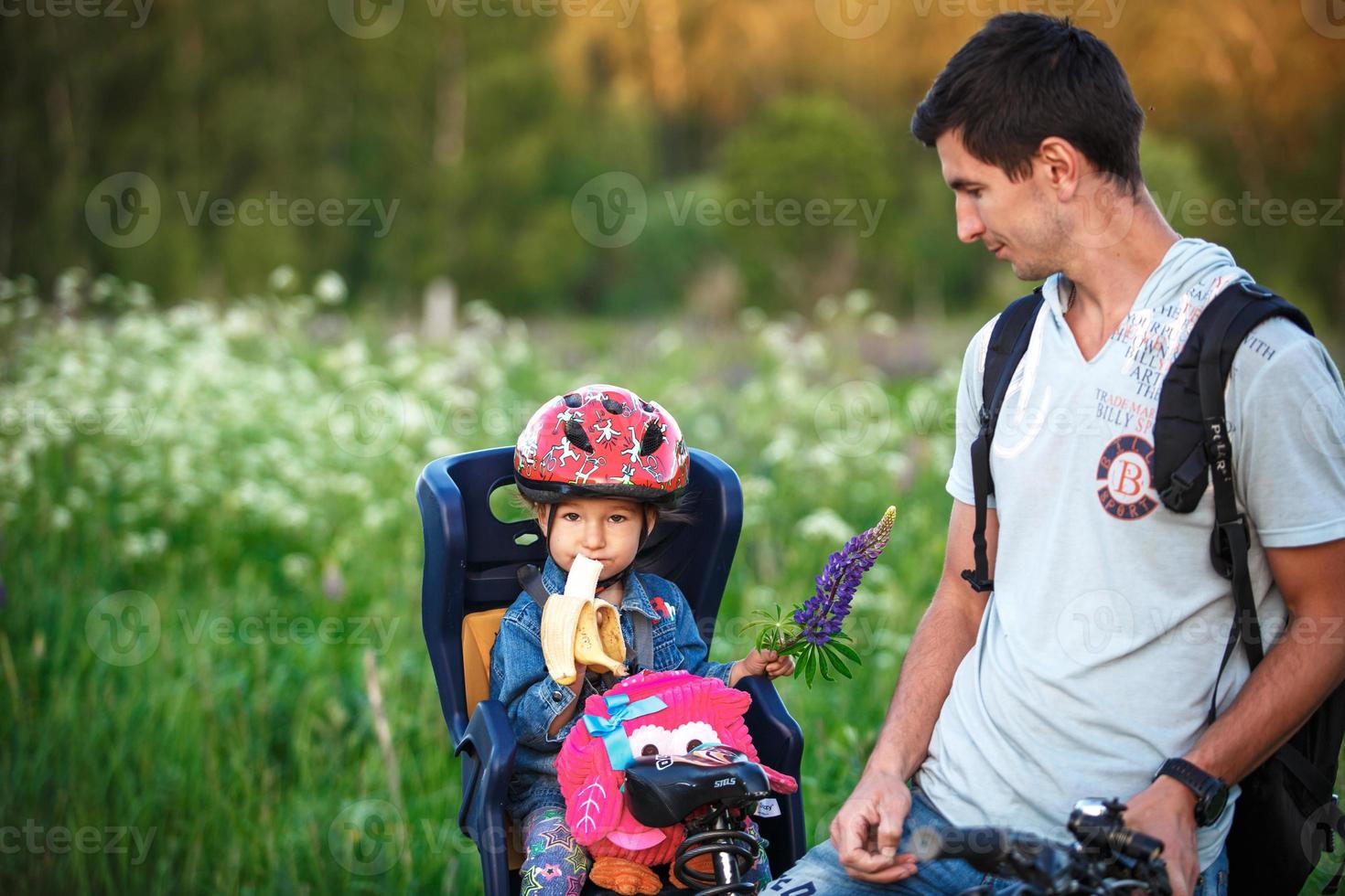 padre con una figlia piccola nel seggiolino di una bicicletta per bambini su una bicicletta per adulti. una ragazza con un casco protettivo, con una banana e uno zaino. passeggiata sportiva in famiglia, sicurezza. Kaluga, Russia, 30 maggio 2018 foto