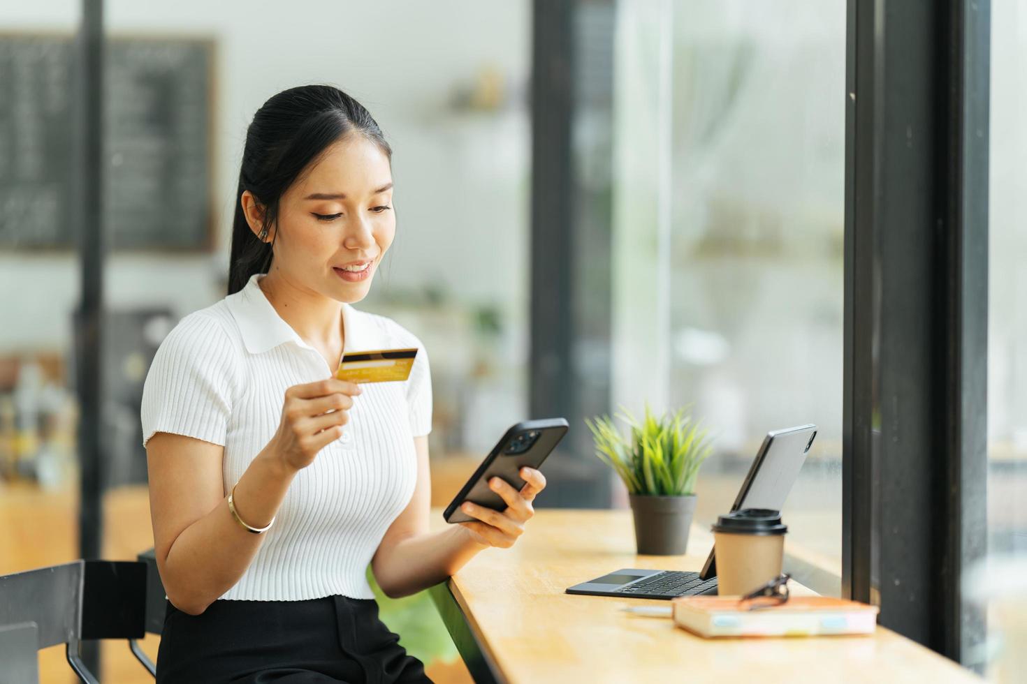 donna sorridente che paga online, utilizza il laptop, tiene in mano una carta di credito in plastica, si siede su una caffetteria, fa acquisti femminili asiatiche, effettua pagamenti sicuri in Internet, naviga nel servizio bancario. foto