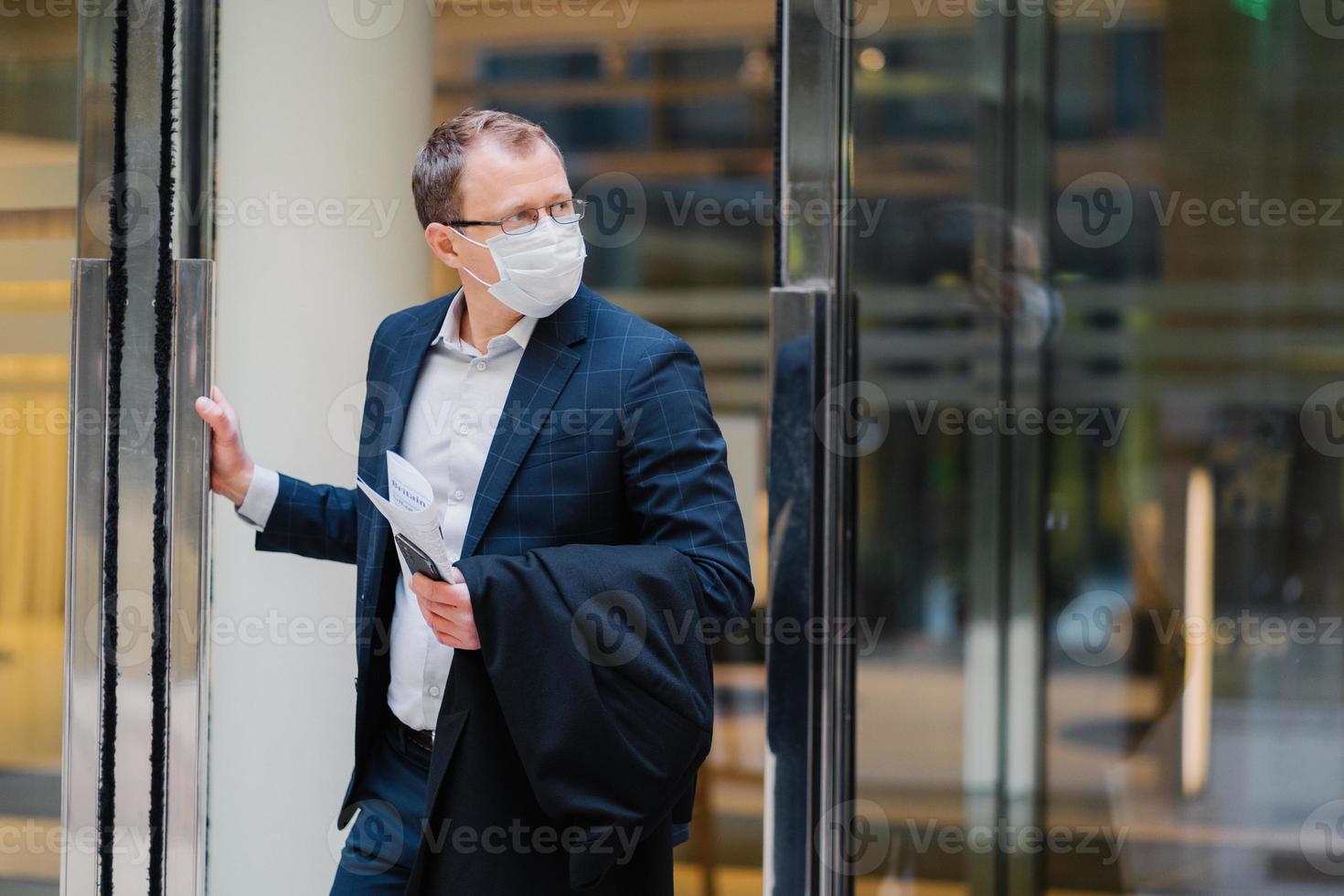 protezione respiratoria, coronavirus, malattie pericolose. un uomo serio si trova sulle porte dell'edificio per uffici, indossa una maschera medica, tiene un cellulare moderno, un giornale per leggere l'articolo sul trattamento del covid-19 foto