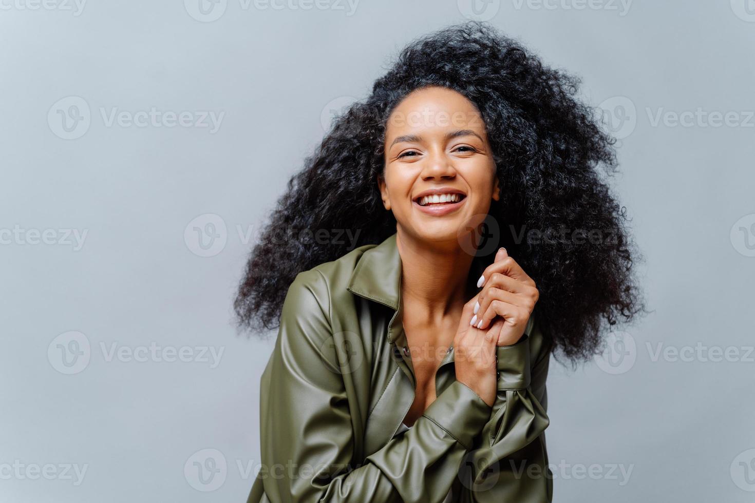 donna gioiosa con la pelle scura, acconciatura afro, tiene le mani unite, sorride sinceramente alla telecamera, mostra denti bianchi, indossa una camicia di pelle, posa su sfondo grigio. persone, emozioni, etnia foto