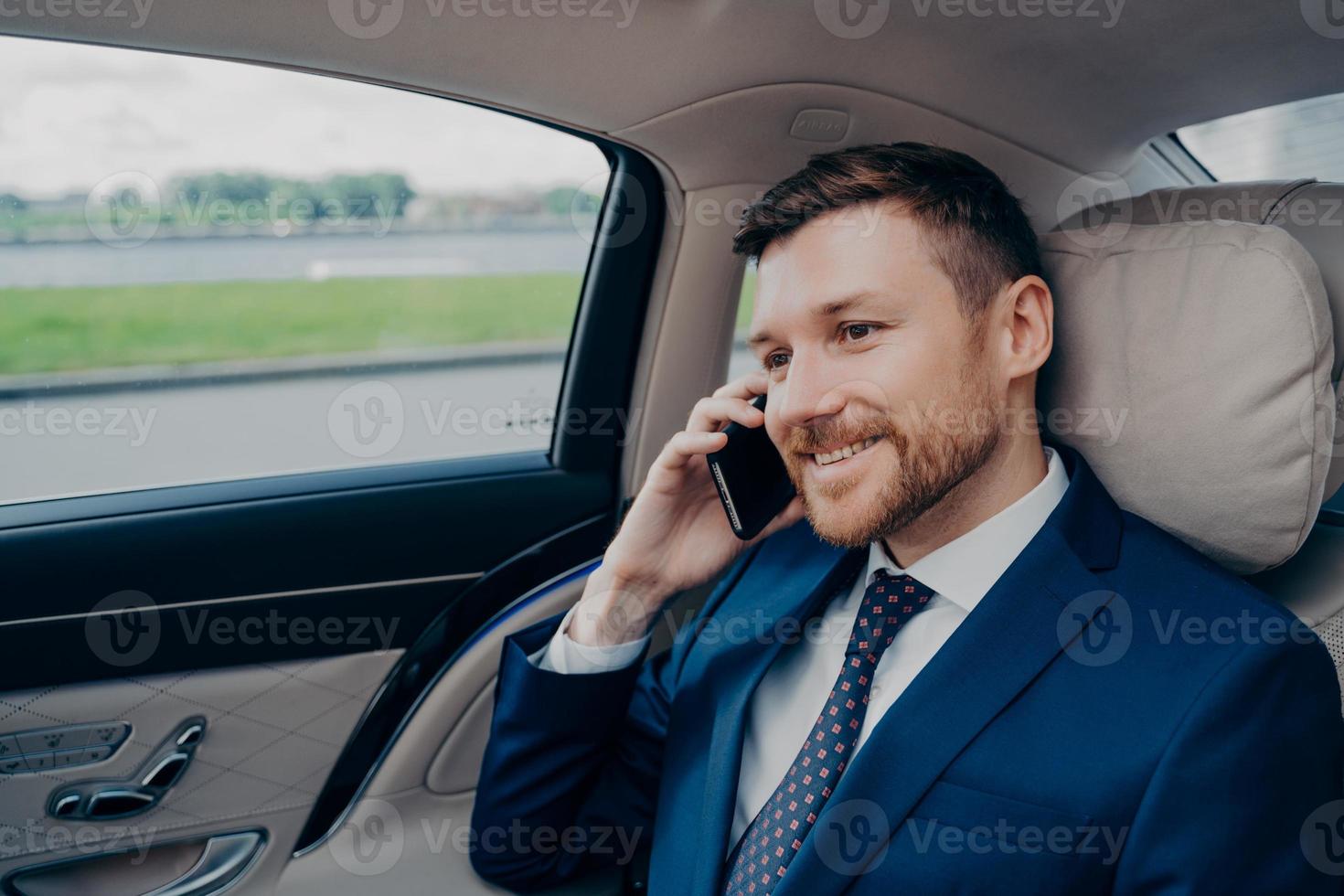 banchiere esperto attraente in abito formale che guida in auto aziendale e parla al cellulare foto