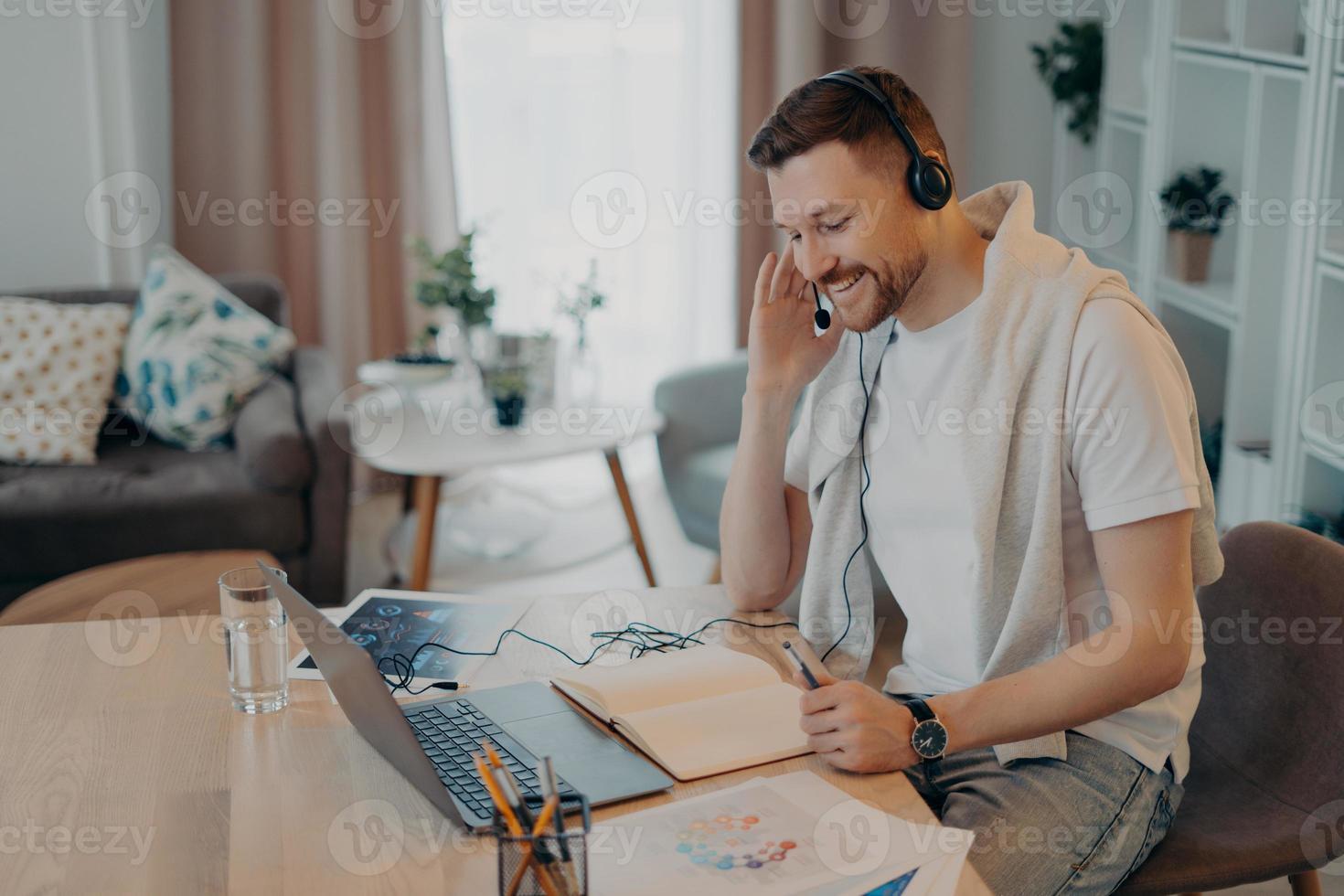 uomo sorridente che usa auricolare e taccuino mentre impara online foto