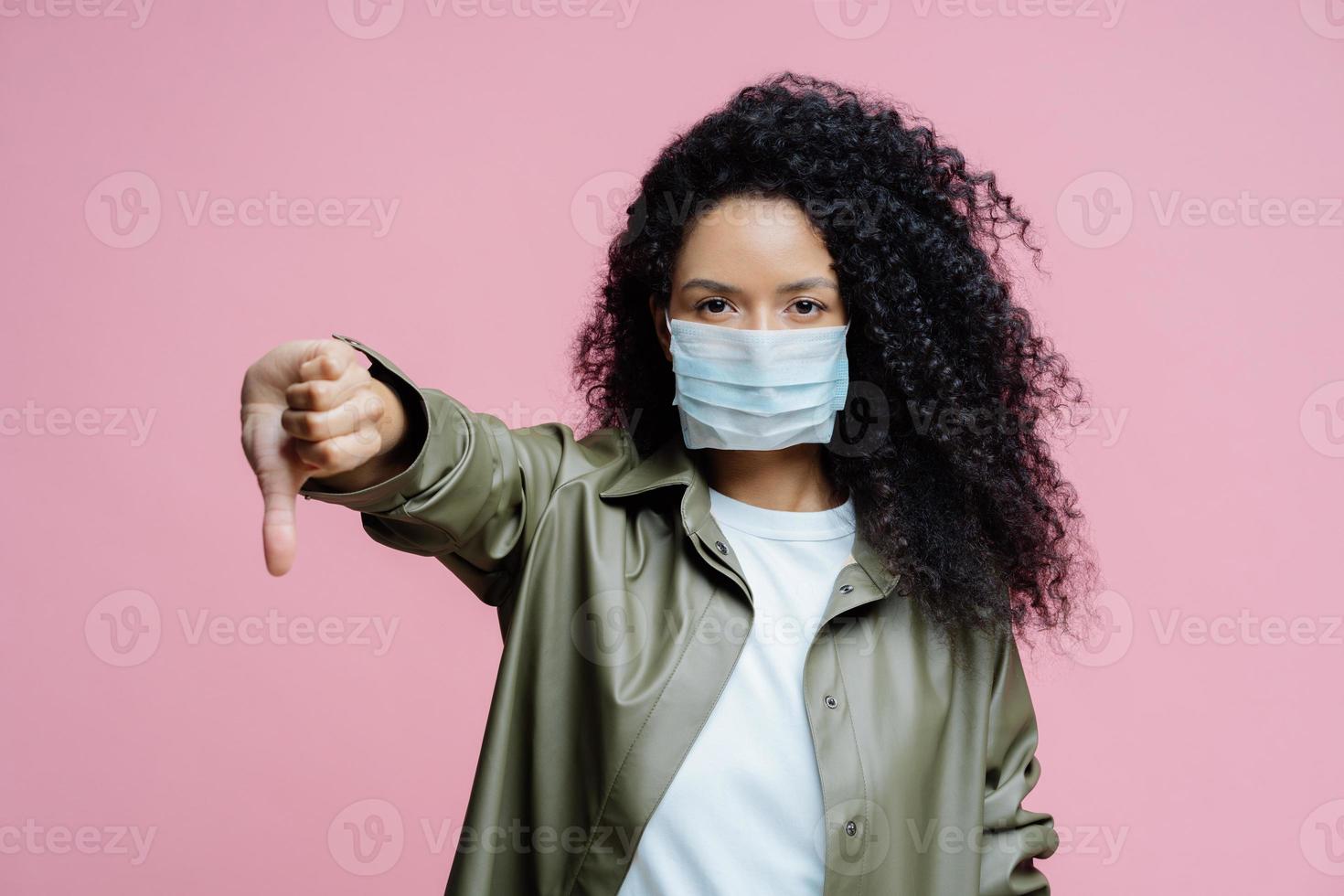 l'immagine di una seria giovane donna dai capelli ricci indossa una maschera medica, tiene il pollice verso il basso, mostra un gesto di antipatia, evita la diffusione del virus corona, si preoccupa della salute. virus pericoloso. concetto medico. foto