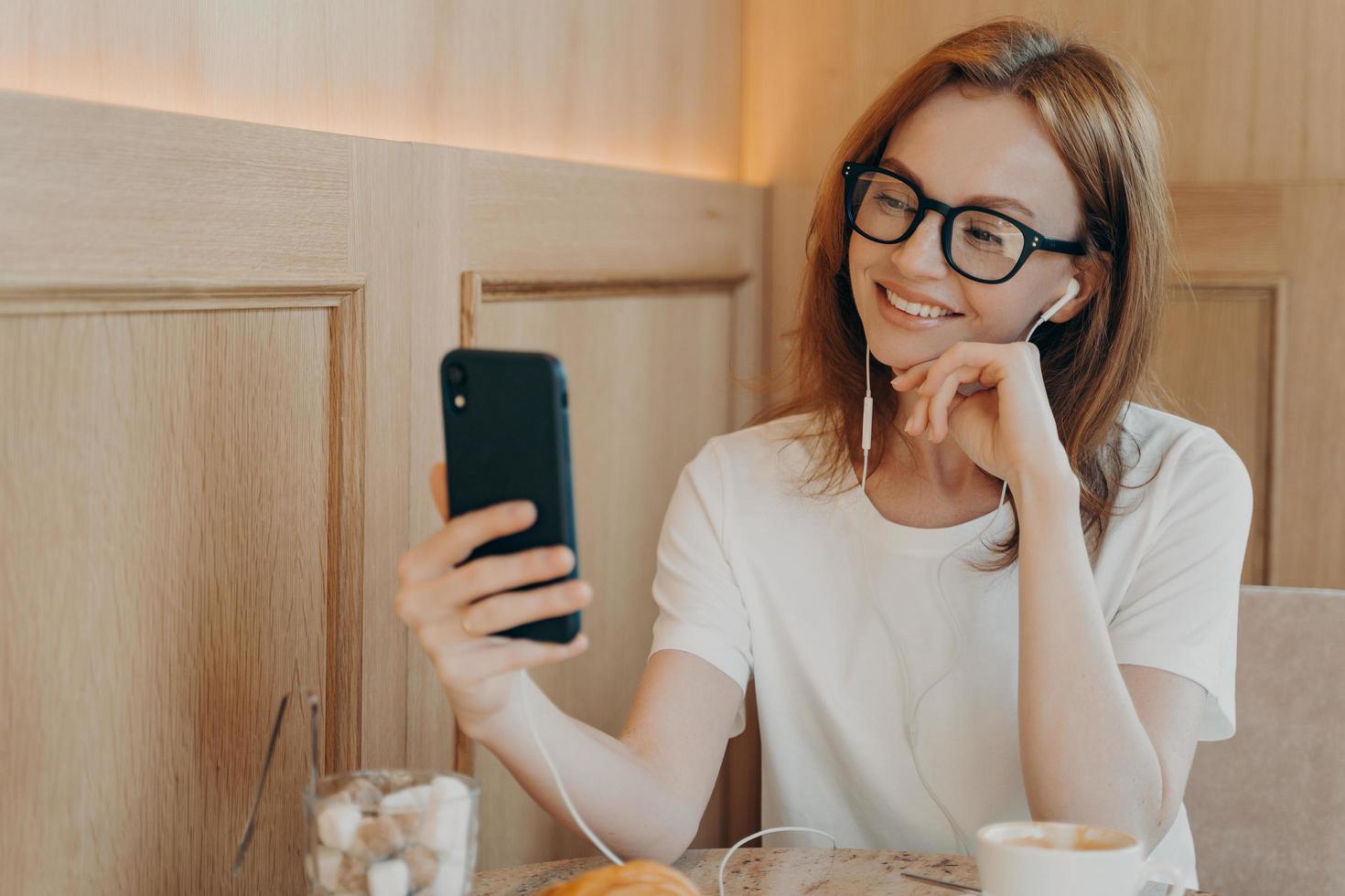 la donna felice sorridente con gli occhiali indossa gli auricolari usa lo smartphone nella caffetteria foto
