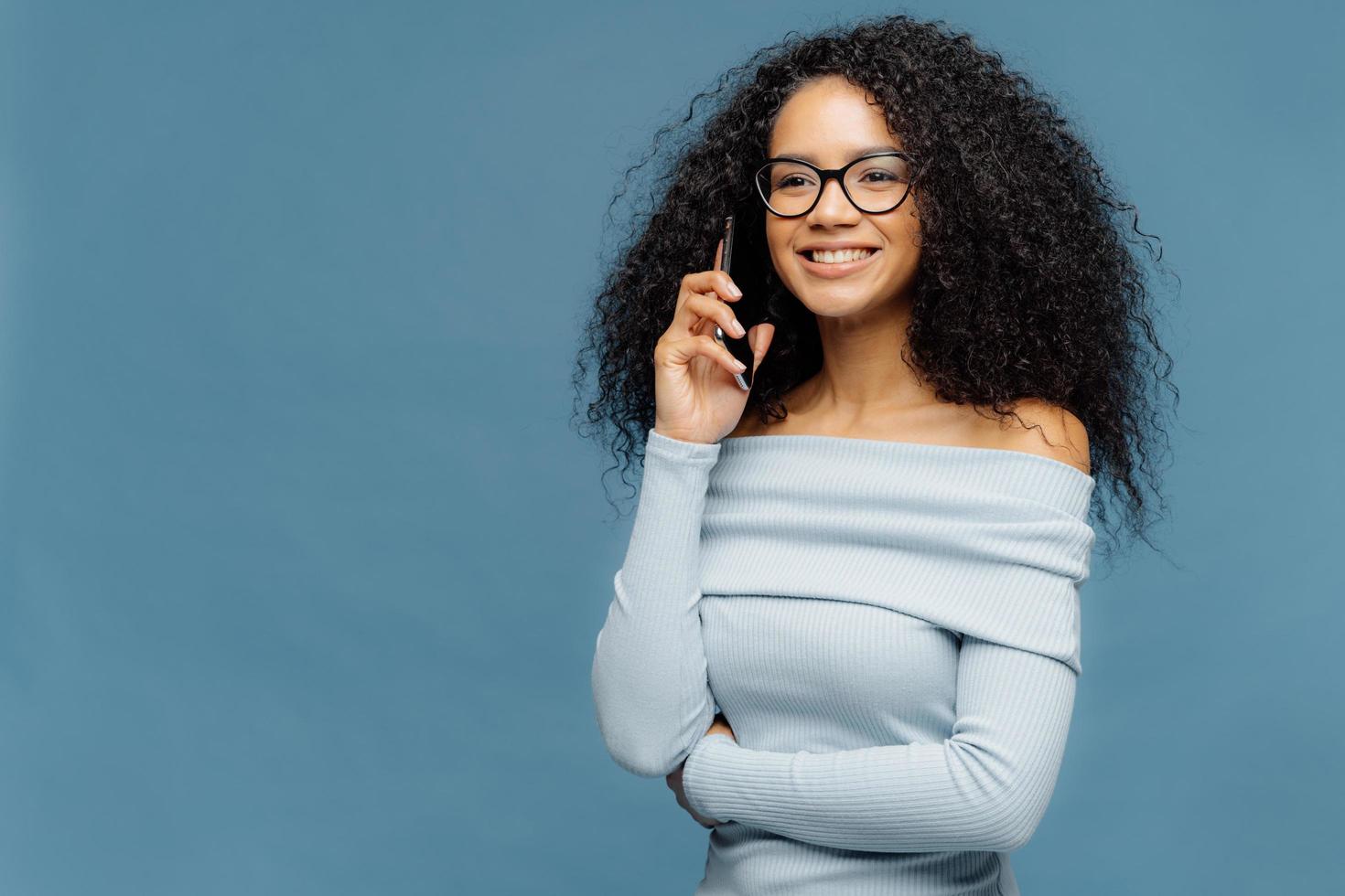 sorridente donna afroamericana felice con i capelli ricci, parla su uno smartphone, discute di alcuni momenti piacevoli dopo la festa, guarda felicemente in lontananza, indossa occhiali e maglione, isolata sul muro blu foto