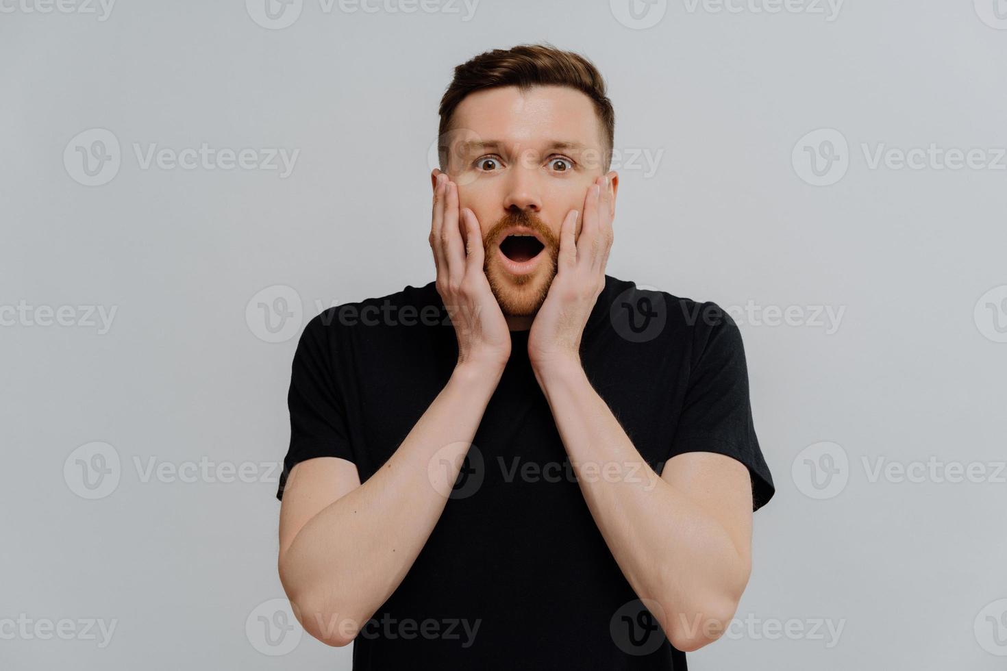 il ritratto di un bell'uomo europeo adulto con la barba tiene le mani sulle guance reagisce su qualcosa di straordinario guarda gli occhi infastiditi vestito con una maglietta nera casual isolata su sfondo blu. omg concetto foto