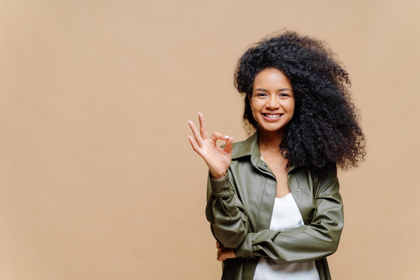 la ripresa in interni di una donna riccia dall'aspetto piacevole ha un sorriso piacevole, fa un gesto ok, segno eccellente, dà approvazione, vestita con una camicia di pelle alla moda, isolata su un muro marrone, spazio vuoto a sinistra foto