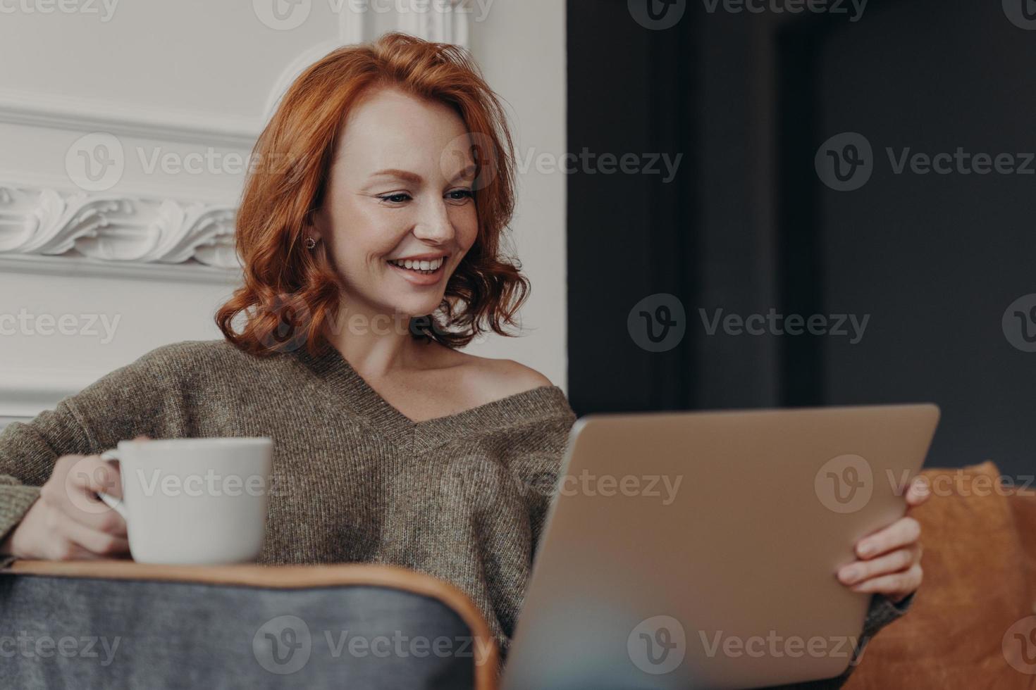 bella donna rossa positiva felice di fare una videochiamata con la migliore amica, condividere notizie tra loro, bere caffè, sedersi in una comoda poltrona, guardare film interessanti, connessa a internet foto