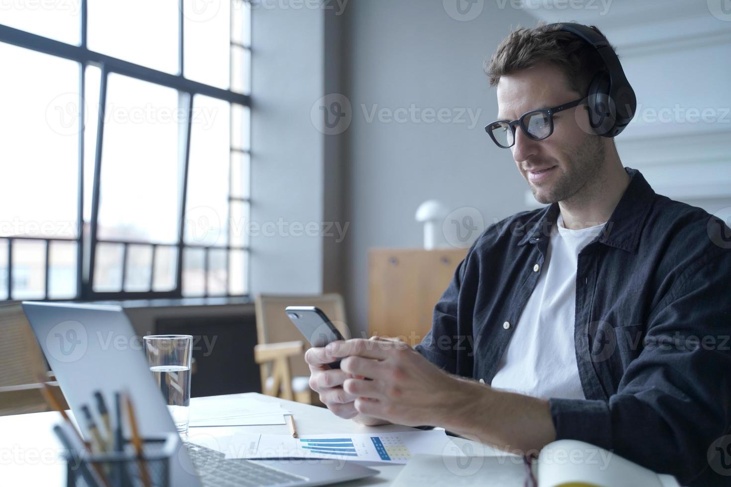 ragazzo freelance tedesco in cuffia seduto in un moderno ufficio a casa tramite laptop mentre si utilizza lo smartphone foto