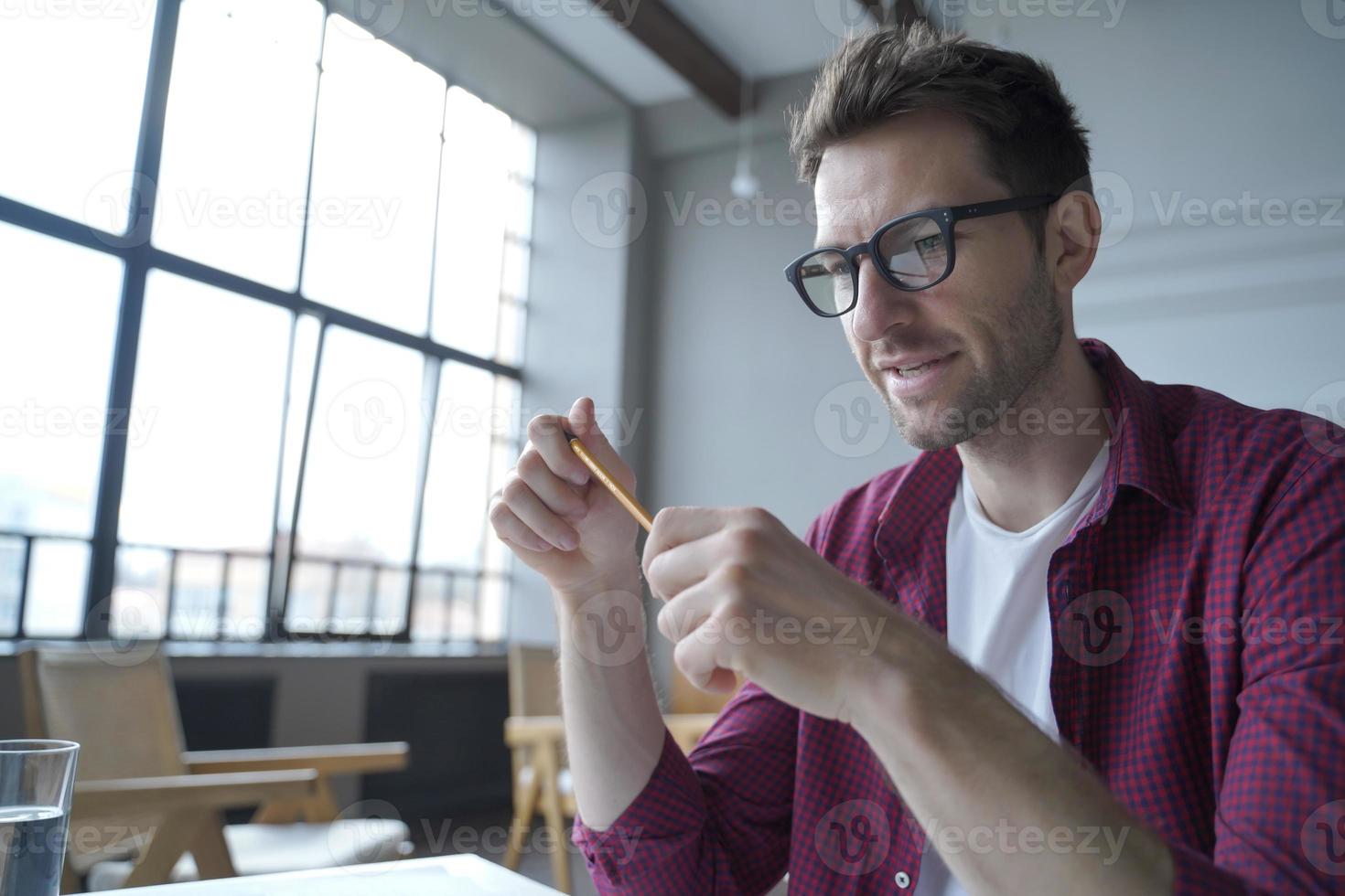 giovane uomo tedesco concentrato con gli occhiali che tiene la matita seduto alla scrivania e dà lezione web online foto