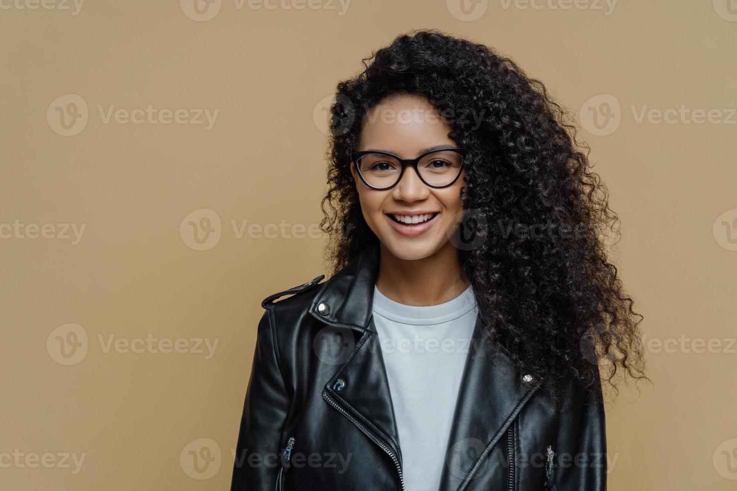 ragazza hipster alla moda con un folto taglio di capelli afro, sorride positivamente, si gode una bella giornata, indossa occhiali e giacca di pelle nera, posa su sfondo marrone. persone, emozioni e concetto di moda foto