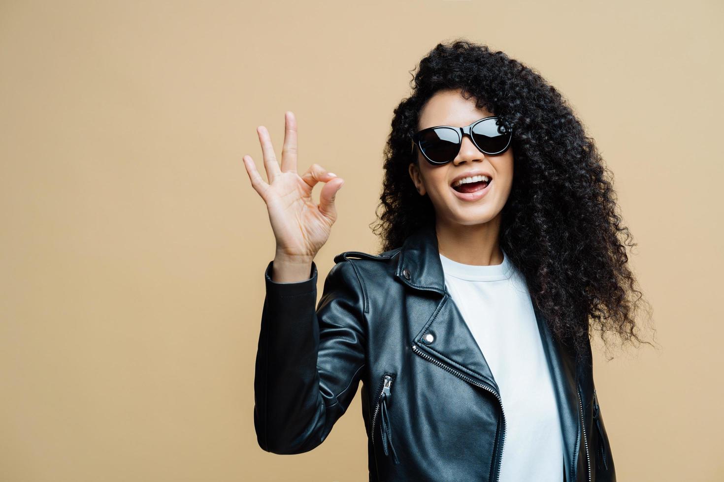 la donna dai capelli ricci alla moda dice che va tutto bene, fa un gesto ok, dice di sì a nuove opportunità, indossa occhiali da sole alla moda e giacca di pelle nera, isolata su sfondo marrone. linguaggio del corpo foto