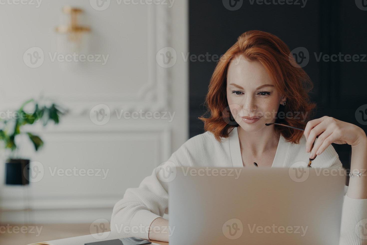 foto di una giornalista seria dai capelli rossi concentrata al computer portatile, cerca informazioni per un articolo sul sito Web di Internet, tiene gli occhiali, indossa un maglione bianco, controlla la posta elettronica, lavora in remoto