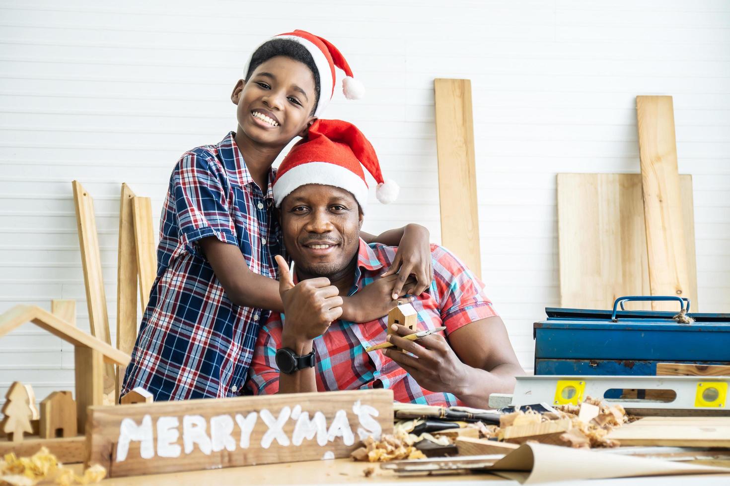 natale felice bambino e padre. allegro figlio afroamericano falegname che abbraccia suo padre mentre si appoggia al tavolo di legno con diversi strumenti di lavoro adagiati su di esso foto