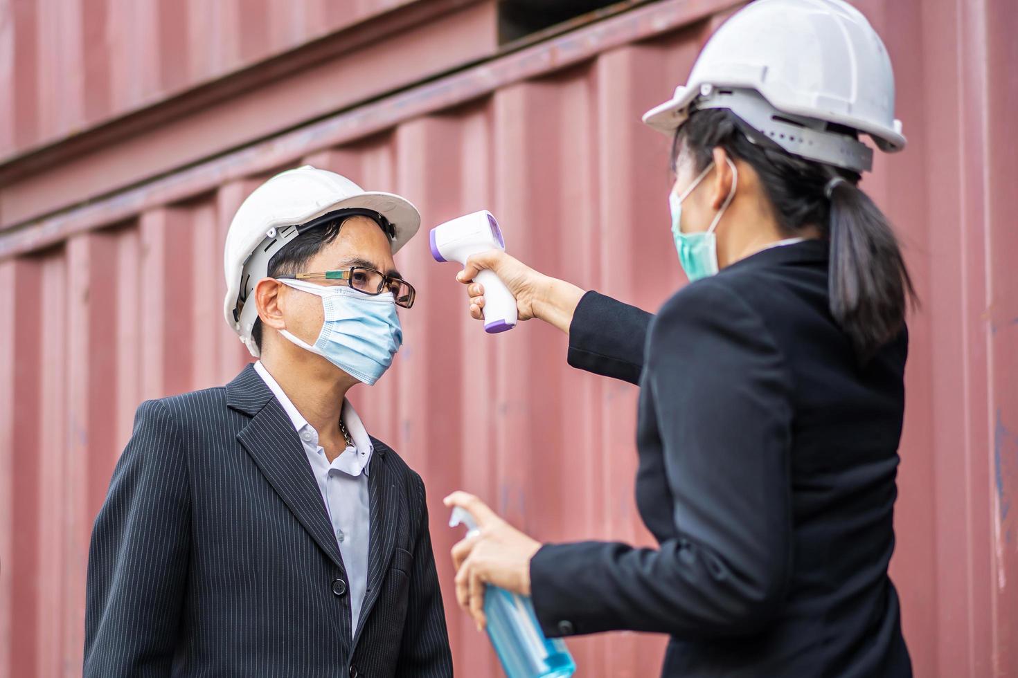 il caposquadra femminile che indossa una maschera misura la temperatura di un caposquadra maschio in abito. si sono fermati e hanno aspettato la misurazione con un termometro a infrarossi senza contatto per prevenire il virus covid foto