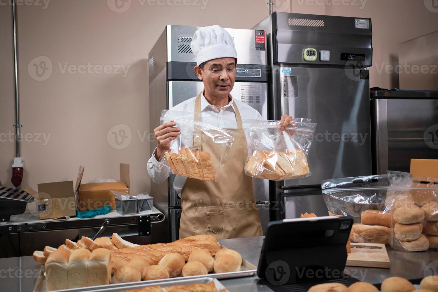 chef maschio asiatico senior in grembiule, blogger di piccole imprese in diretta streaming tramite tecnologia di applicazione mobile, promozione online di vendita di prodotti da forno, pane fresco, pasticcini e cucina commerciale in cucina. foto