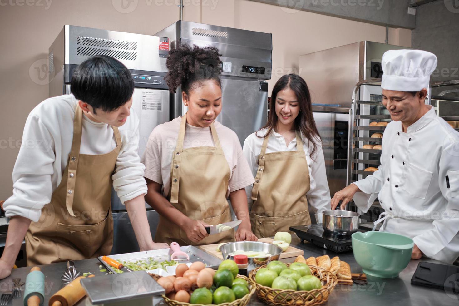 corso di cucina per hobby, chef maschio senior in uniforme da cuoco insegna ai giovani studenti delle lezioni di cucina a sbucciare e tagliare le mele, ingredienti per pasticcini, torte di frutta nella cucina in acciaio inossidabile del ristorante. foto