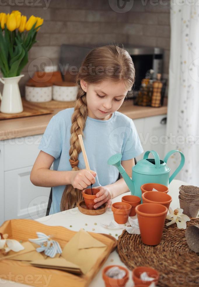 bambina seduta al tavolo di casa, seminando semi in vasi di fiori. foto